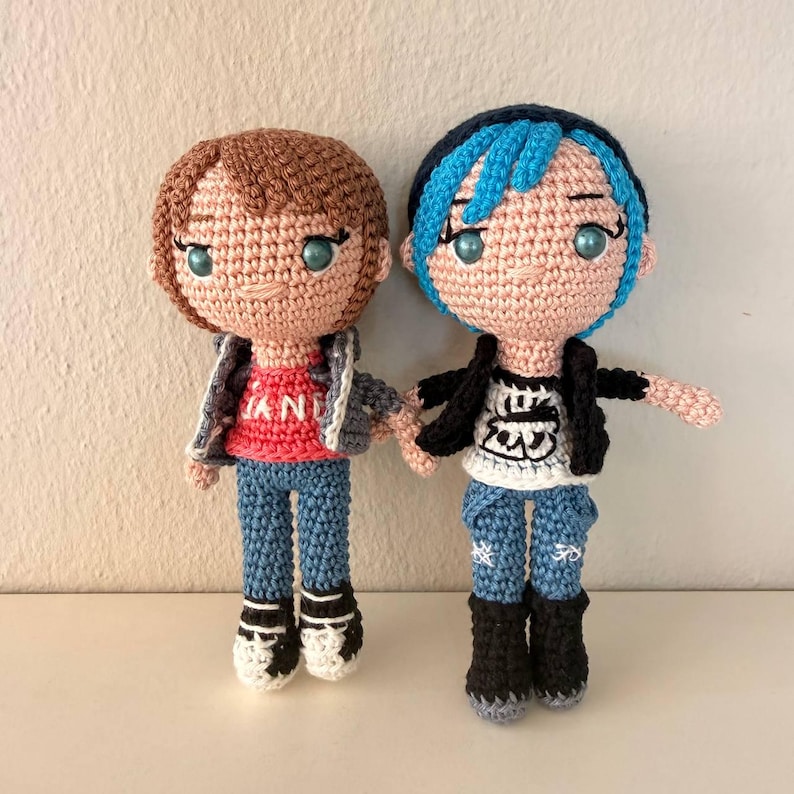 May include: Two handmade crochet dolls. One doll has brown hair, a red shirt with white text, a gray jacket, blue jeans, and black and white shoes. The other doll has blue hair, a black jacket, a white shirt with a graphic, blue jeans, and black boots.