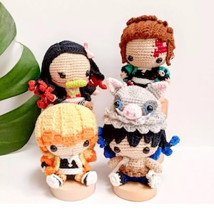 May include: Four crocheted anime character dolls. The dolls feature detailed yarn work in various colors, including black, orange, brown, and blue. Each doll is seated on a small wooden base, showcasing their unique outfits and hairstyles.