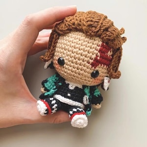 May include: A crocheted doll with brown hair and a facial scar. The doll is dressed in a black and green outfit with white and red details. It has black button eyes. The doll is held in a hand.