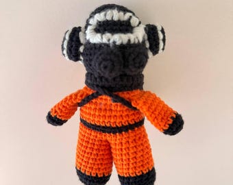 May include: A crochet doll shaped like an astronaut wearing a bright orange jumpsuit with black trim at the waist, wrists, and ankles. The head features a black helmet with a white circular faceplate border and two small black circular ears.