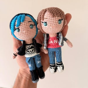 May include: Two handmade crochet dolls, each with a unique style. One doll has blue hair, a black jacket, and a graphic tee. The other doll has brown hair, a gray jacket, and a red shirt. Both dolls wear blue jeans and black boots.