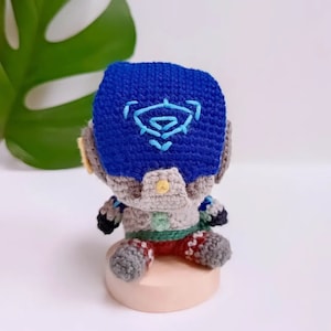 Könnte beinhalten: Ein gehäkeltes Plüschtier eines Robotercharakters, der einen blauen Helm mit einem gestickten, stilisierten Symbol trägt. Der Roboter sitzt auf einer kleinen, hellbraunen Basis.
