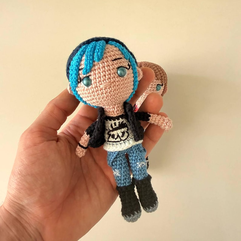 May include: Crocheted doll with blue hair, wearing a black jacket, white t-shirt, and blue jeans. The doll has black boots and blue eyes. A second doll is partially visible in the background.