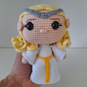 May include: A crocheted doll with blonde hair, a white dress, and a yellow sash. The doll has a gray headband and black eyes.
