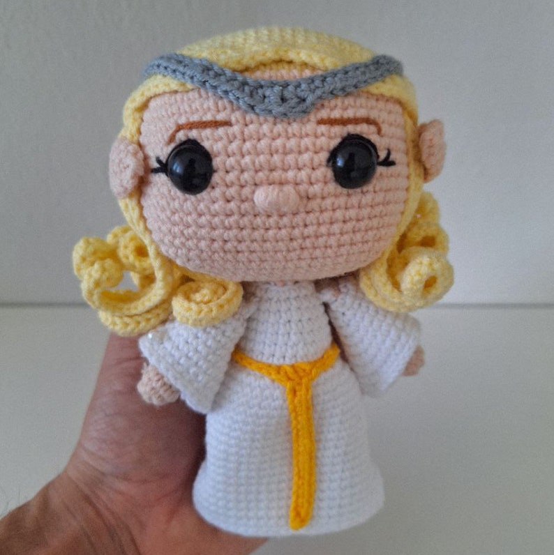 May include: A crocheted doll with blonde hair, a white dress, and a yellow sash. The doll has a gray headband and black eyes.