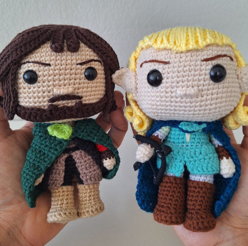 May include: Two crocheted plush toys, one with brown hair and a green cloak, the other with blonde hair and a blue cloak.