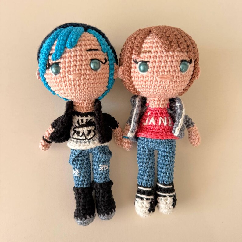 May include: Two handmade crochet dolls. One doll has blue hair, a black jacket, and blue jeans. The other doll has brown hair, a gray jacket, and blue jeans. Both dolls have light skin and are wearing sneakers or boots.