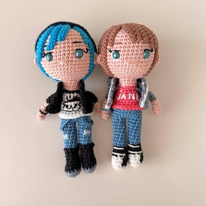 May include: Two handmade crochet dolls. One doll has blue hair, a black jacket, and blue jeans. The other doll has brown hair, a gray jacket, and blue jeans. Both dolls have light skin and blue eyes. The dolls are standing side by side.