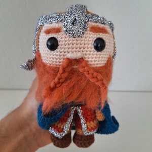 May include: A crocheted plush toy of a dwarf with a red beard, silver helmet, and blue and red clothing.