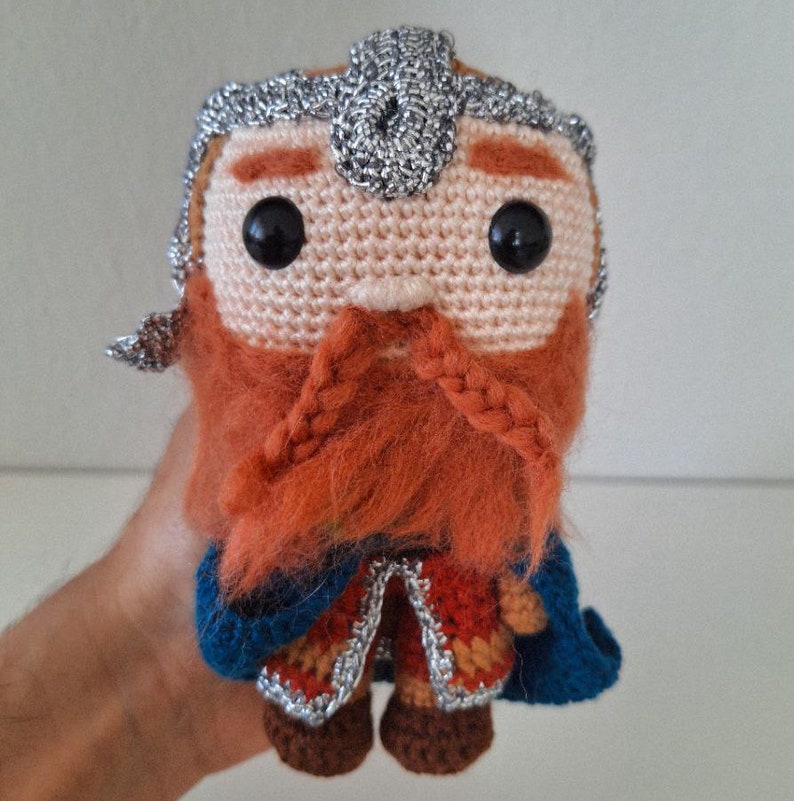 May include: A crocheted plush toy of a dwarf with a red beard, silver helmet, and blue and red clothing.