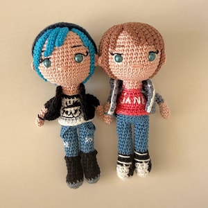 May include: Two handmade crochet dolls. One doll has blue hair, a black jacket, and blue jeans. The other doll has brown hair, a gray jacket, a red shirt, and blue jeans. Both dolls have light blue eyes and are made of yarn.