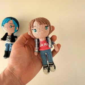 May include: Two crochet dolls with yarn hair and embroidered facial features. One doll has blue hair, a black jacket, and blue pants. The other doll has brown hair, a red shirt with the text JANE, a grey jacket, and blue pants with white shoes.