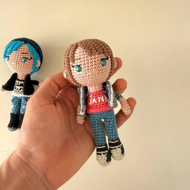 May include: Two crochet dolls with yarn hair and embroidered facial features. One doll has blue hair, a black jacket, and blue pants. The other doll has brown hair, a red shirt with the text JANE, a grey jacket, and blue pants with white shoes.