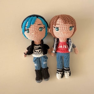 May include: Two handmade crochet dolls with yarn hair and embroidered features. The left doll has blue hair, a black vest, white graphic shirt, blue pants, and black boots. The right doll has brown hair, a gray jacket, red shirt with text, blue pants, and white sneakers.