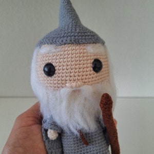 May include: A crocheted gray and white wizard doll with a brown staff. The doll has a pointed hat, a white beard, and black eyes.