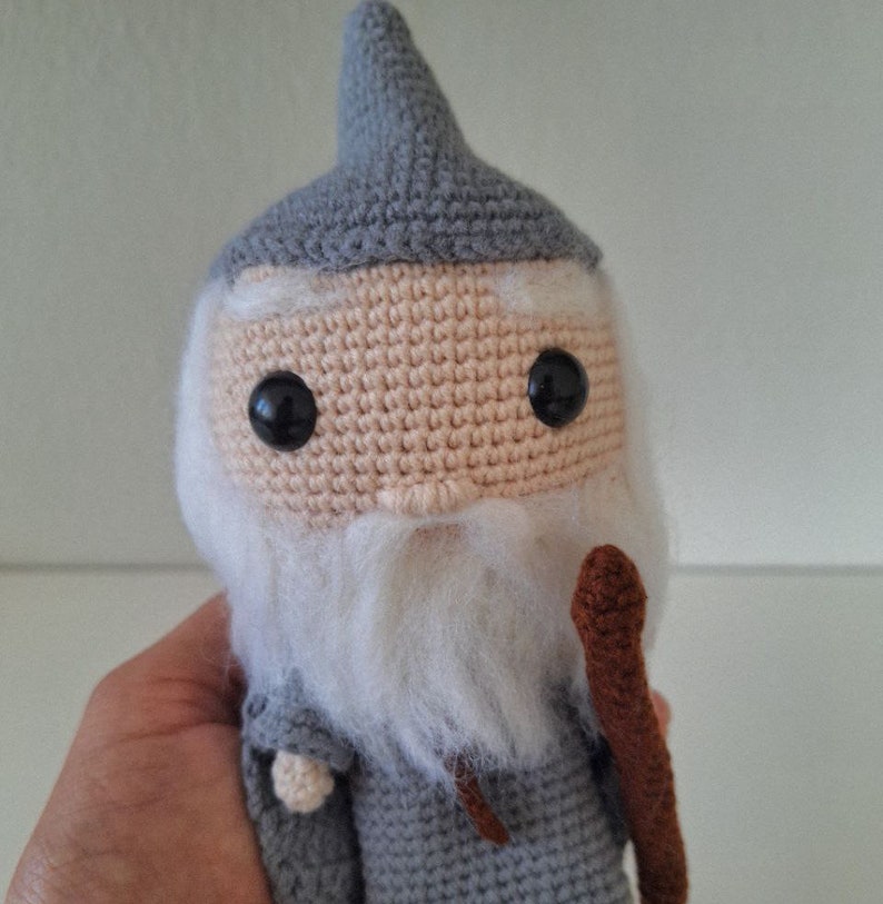 May include: A crocheted gray and white wizard doll with a brown staff. The doll has a pointed hat, a white beard, and black eyes.