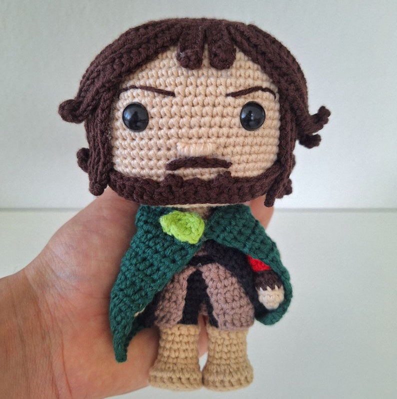 May include: Crocheted amigurumi toy of a character with brown hair, a green cloak, and a brown beard. The toy has black eyes and a serious expression.