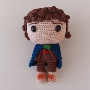 May include: Crocheted amigurumi doll with brown hair, a blue cape, and brown pants. The doll has a round face with brown eyes and a small, green leaf on its chest.