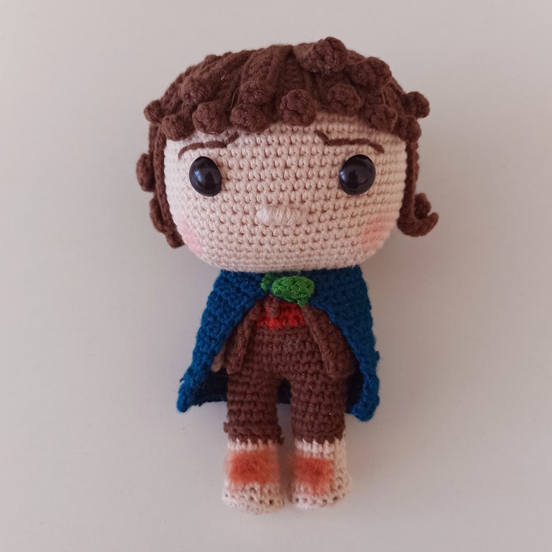 May include: Crocheted amigurumi doll with brown hair, a blue cape, and brown pants. The doll has a round face with brown eyes and a small, green leaf on its chest.