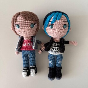 May include: Two handmade crochet dolls. One doll has brown hair, a red shirt with the word "LAND" on it, a denim jacket, and blue jeans. The other doll has blue and black hair, a black jacket, a white shirt with a graphic, and blue jeans.