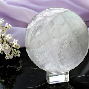 May include: A clear crystal ball on a clear square base. The sphere has internal patterns and sits on a reflective black surface. White flowers and a lavender fabric are in the background.