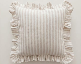 May include: A square throw pillow featuring a vertical striped pattern in beige and white. The pillow is finished with a gathered ruffle trim along all four edges, constructed from the same striped fabric as the main body of the cushion.