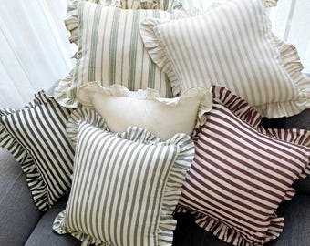 May include: A collection of square throw pillows featuring ruffled edges. The pillows display various striped patterns in shades of green, beige, and dark red, alongside one solid off-white pillow, all arranged together in a group.