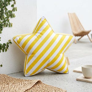 May include: A star-shaped throw cushion featuring a pattern of diagonal yellow and white stripes. The cushion has white piping along its edges and a small beige fabric tag on the side with the text Hello Mello printed in a dark colour.