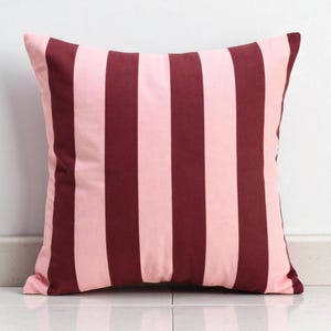 May include: Square decorative pillow with a striped pattern. The pillow features alternating vertical stripes of light pink and dark burgundy. The pillow is resting on a white tiled surface.