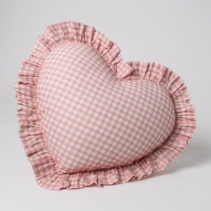 Könnte beinhalten: Ein herzförmiges Kissen mit rosa-weißem Vichy-Muster. Das Kissen hat einen gerüschten Rand aus passendem rosa Stoff. Es ist ein dekorativer Artikel, wahrscheinlich für ein Bett oder Sofa.