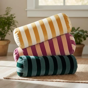 May include: Three striped bolster pillows in shades of yellow and white, purple and peach, and dark green. The pillows are stacked on top of each other on a woven rug. The pillows are cylindrical in shape and appear to be made of a soft, velvety material.