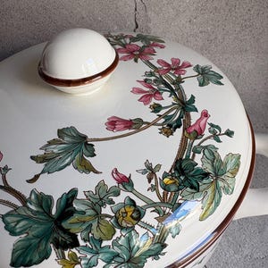 Peut inclure: Un plat de service en c&eacute;ramique blanche avec un couvercle et une poign&eacute;e. Le couvercle comporte un bouton blanc avec un bord marron. Le plat est d&eacute;cor&eacute; d'un motif floral rose, vert et marron. Le plat est probablement utilis&eacute; pour servir des aliments.