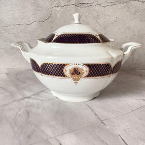May include: White porcelain soup tureen with a lid, featuring a dark blue and gold geometric pattern. The tureen has two handles and a decorative gold emblem. A classic piece for serving soups or stews.