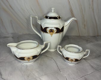 Könnte beinhalten: Ein weißes Porzellan-Teeservice mit Teekanne, Sahnekännchen und Zuckerdose. Das Set hat ein schwarzes und goldenes geometrisches Design. Die Teekanne hat einen Henkel und einen Ausguss.