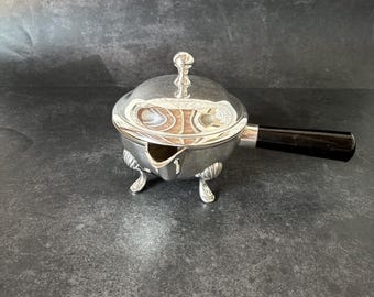 Peut inclure: Un saucier en argent avec un couvercle et une poignée noire. Le saucier a trois pieds et un bec verseur. Le saucier est sur une surface gris foncé.
