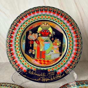 Può includere: Un set di tre piatti in porcellana blu e oro con disegni colorati. Il piatto centrale presenta un re con una corona e un uccello, circondato da un bordo colorato. Il piatto è firmato "Weihnachten 1992" e "Bjorn Wiinblad".