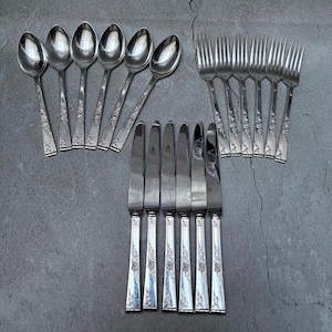 Peut inclure: Un ensemble de couverts anciens, comprenant des cuillères, des fourchettes et des couteaux, disposés sur une surface grise. Les couverts présentent un motif floral sur les manches. Les cuillères, fourchettes et couteaux sont en métal brillant de couleur argent.