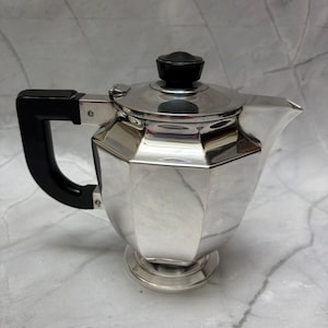 Peut inclure: Une théière en métal argenté poli avec un corps octogonal, un bec court et une poignée noire. Le couvercle est surmonté d'un petit bouton noir et la base est circulaire. La théière présente une surface réfléchissante et un design géométrique.