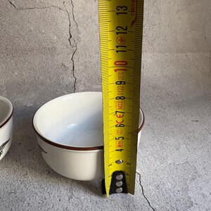 Peut inclure: Un bol en c&eacute;ramique blanc avec un bord marron, d'environ 10 cm de diam&egrave;tre. Un m&egrave;tre ruban jaune est positionn&eacute; &agrave; c&ocirc;t&eacute; du bol, indiquant la mesure en centim&egrave;tres. Le bol a un design simple et &eacute;pur&eacute;.