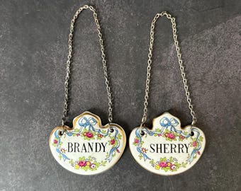 May include: Two vintage porcelain bottle labels with silver chains. The labels are white with blue and gold trim and floral designs. One label says "BRANDY" and the other says "SHERRY".
