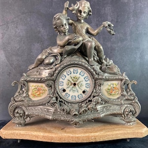 Könnte beinhalten: Eine silberfarbene Kaminuhr mit einem Porzellanzifferblatt und Cherubfiguren. Die Uhr ist mit floralen Details verziert und hat einen Marmorsockel.