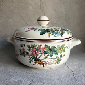 Peut inclure: Un saladier en c&eacute;ramique cr&egrave;me avec couvercle. Le bol pr&eacute;sente un motif floral avec des fleurs roses, des feuilles vertes et des racines brunes. Le couvercle a un bouton rond et un bord marron. Le bol a deux poign&eacute;es.