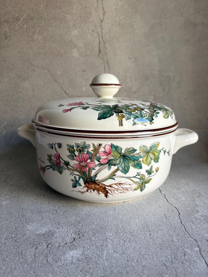 Peut inclure: Un saladier en c&eacute;ramique cr&egrave;me avec couvercle. Le bol pr&eacute;sente un motif floral avec des fleurs roses, des feuilles vertes et des racines brunes. Le couvercle a un bouton rond et un bord marron. Le bol a deux poign&eacute;es.