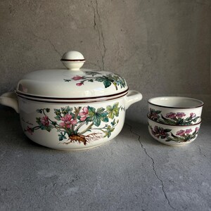 Peut inclure: Une soupi&egrave;re en c&eacute;ramique cr&egrave;me avec un couvercle, deux poign&eacute;es et un motif floral avec des fleurs roses et des feuilles vertes. Deux petits bols assortis sont empil&eacute;s &agrave; droite. La soupi&egrave;re et les bols ont une bordure marron fonc&eacute;.