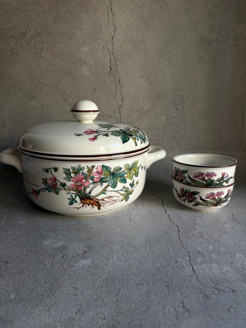 Peut inclure: Une soupi&egrave;re en c&eacute;ramique cr&egrave;me avec un couvercle, deux poign&eacute;es et un motif floral avec des fleurs roses et des feuilles vertes. Deux petits bols assortis sont empil&eacute;s &agrave; droite. La soupi&egrave;re et les bols ont une bordure marron fonc&eacute;.