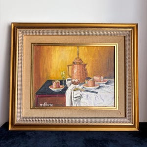 May include: An oil painting in a gold frame depicting a still life with a tall copper-coloured coffee pot, two small pink cups on saucers, a clear glass, and a dark-handled utensil arranged on a white cloth-covered table against a yellow wall.
