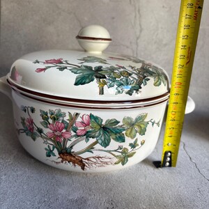 Peut inclure: Un plat &agrave; casserole en c&eacute;ramique blanche avec un couvercle, orn&eacute; d'un motif floral avec des fleurs roses et des feuilles vertes. Le plat a un bord et des poign&eacute;es marron. Le couvercle a un bouton rond. Le plat mesure environ 20 cm de diam&egrave;tre.