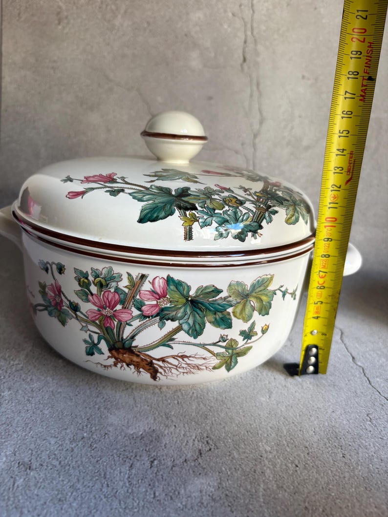Peut inclure: Un plat &agrave; casserole en c&eacute;ramique blanche avec un couvercle, orn&eacute; d'un motif floral avec des fleurs roses et des feuilles vertes. Le plat a un bord et des poign&eacute;es marron. Le couvercle a un bouton rond. Le plat mesure environ 20 cm de diam&egrave;tre.