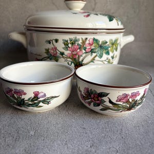 Peut inclure: Un plat de service en c&eacute;ramique blanche avec un couvercle et deux bols assortis. L'ensemble pr&eacute;sente un motif floral avec des fleurs roses et violettes, des feuilles vertes et une bordure marron. Le plat a deux poign&eacute;es et un bouton rond sur le couvercle.