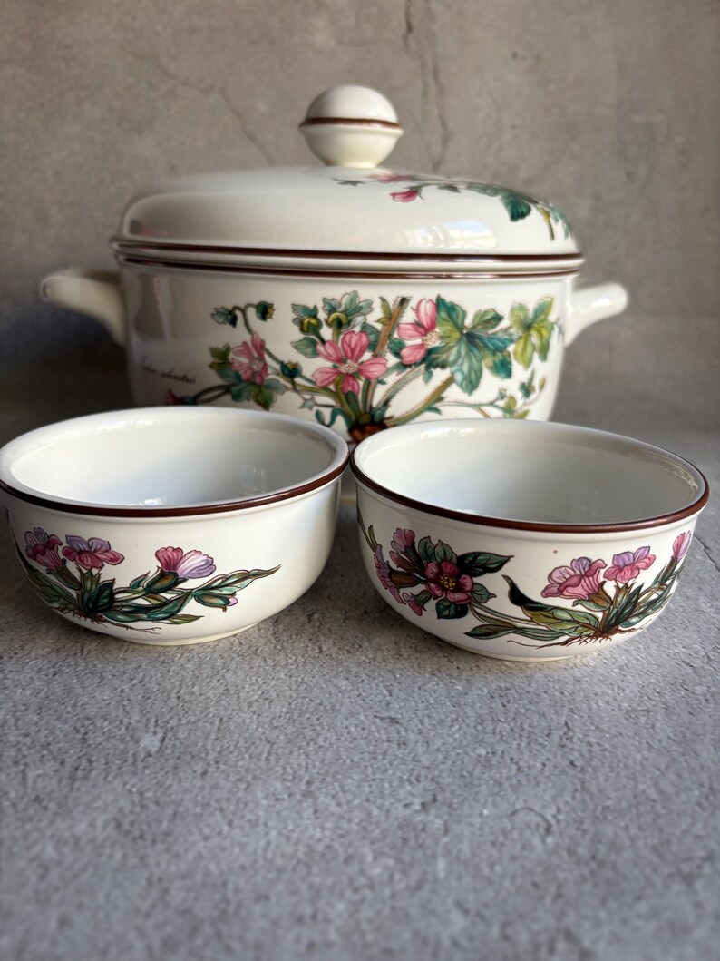 Peut inclure: Un plat de service en c&eacute;ramique blanche avec un couvercle et deux bols assortis. L'ensemble pr&eacute;sente un motif floral avec des fleurs roses et violettes, des feuilles vertes et une bordure marron. Le plat a deux poign&eacute;es et un bouton rond sur le couvercle.